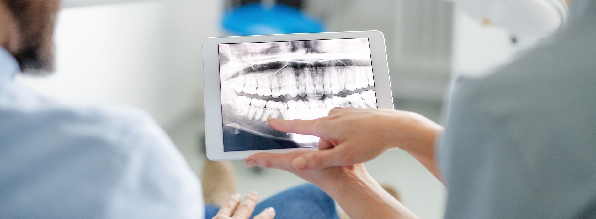 A person holding a tablet displaying an X-ray image while another person looks on, both are situated in a dental office setting.