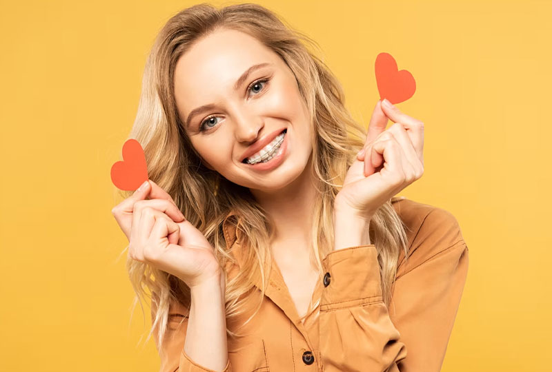 The image features a young woman with blonde hair smiling at the camera, holding up two red heart-shaped objects, possibly candies, with one hand while wearing a brown top and standing against a yellow background.