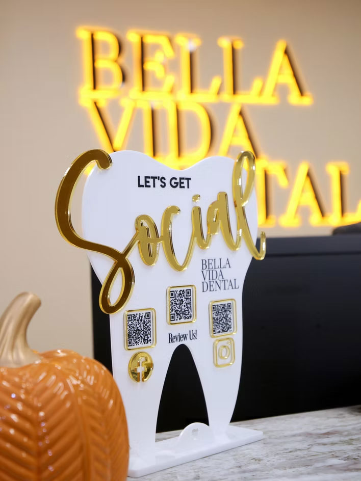 The image features a dental office sign with a tooth-shaped sign that reads Bella Vida Dental and a social media QR code, placed on a desk with a Halloween pumpkin.