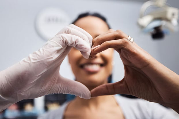 The image shows two people holding their hands together in front of their chests, forming a heart shape, against a background that appears to be an indoor setting with medical equipment visible.