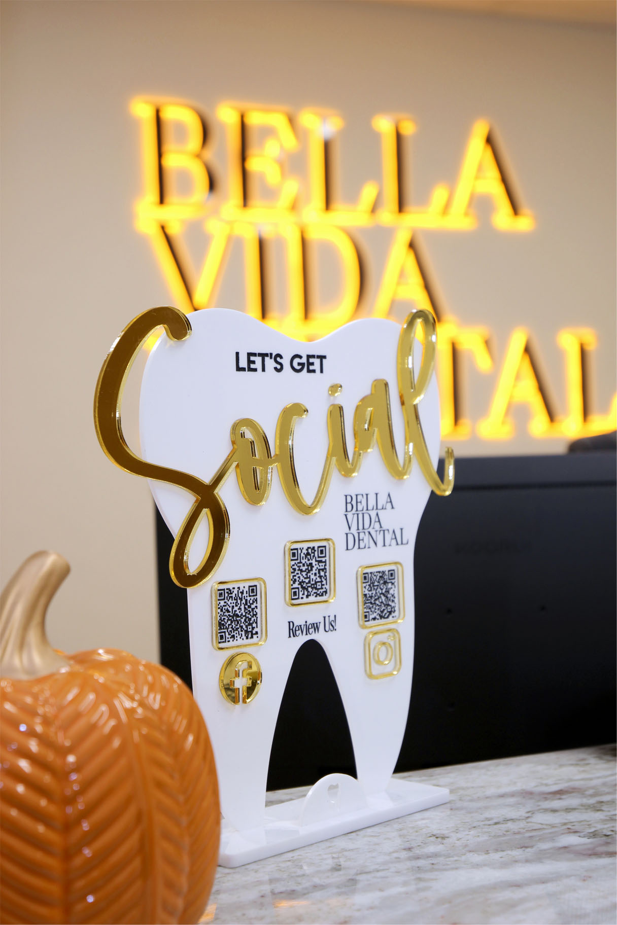 The image shows a sign with the name Bella Vida Social accompanied by a toothbrush-shaped sign that says Let s get social and a QR code, placed on a countertop next to a pumpkin.
