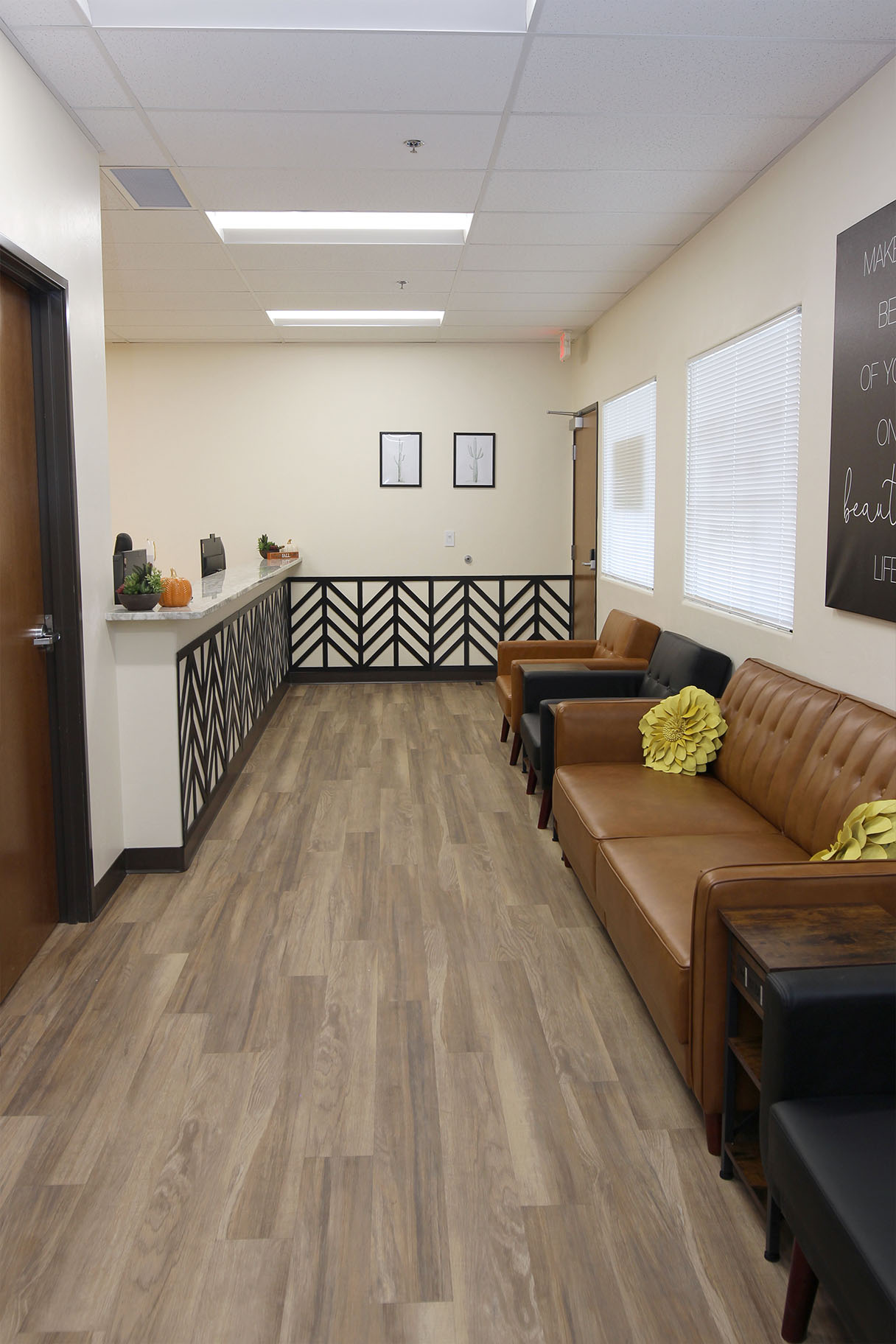 The image depicts an interior hallway with a wooden floor and walls, featuring two couches on either side, a reception desk, a whiteboard, and a door at the end of the hallway.