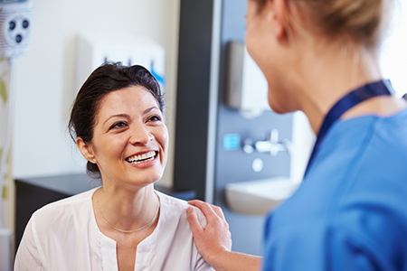 The image shows two individuals, presumably in a medical setting, with one person standing and smiling while another appears to be seated and engaged in conversation.