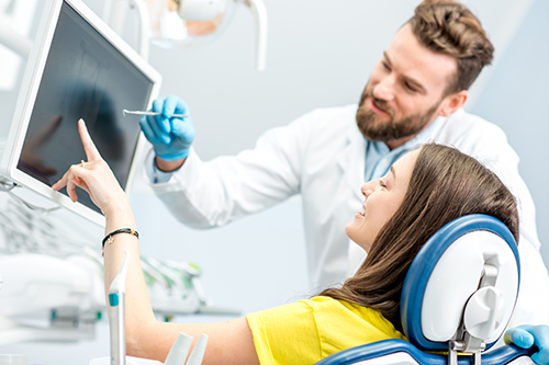 A man in a white coat stands next to a woman seated on a dental chair, both looking at a computer screen they are in a dental office setting.